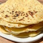 A stack of freshly made flatbreads on a white plate. The flatbreads have golden brown spots and are placed on a wooden table under sunlight.