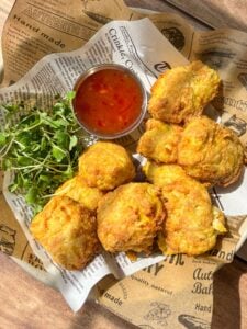 Plate of golden-brown fried snacks served on newspaper-style paper with a side of fresh greens and a small metal cup of orange dipping sauce, all placed on a wooden table.