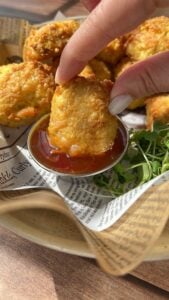 A hand dips a golden, crispy nugget into a small cup of reddish sauce, with more nuggets and some fresh greens on a tray lined with printed parchment paper.