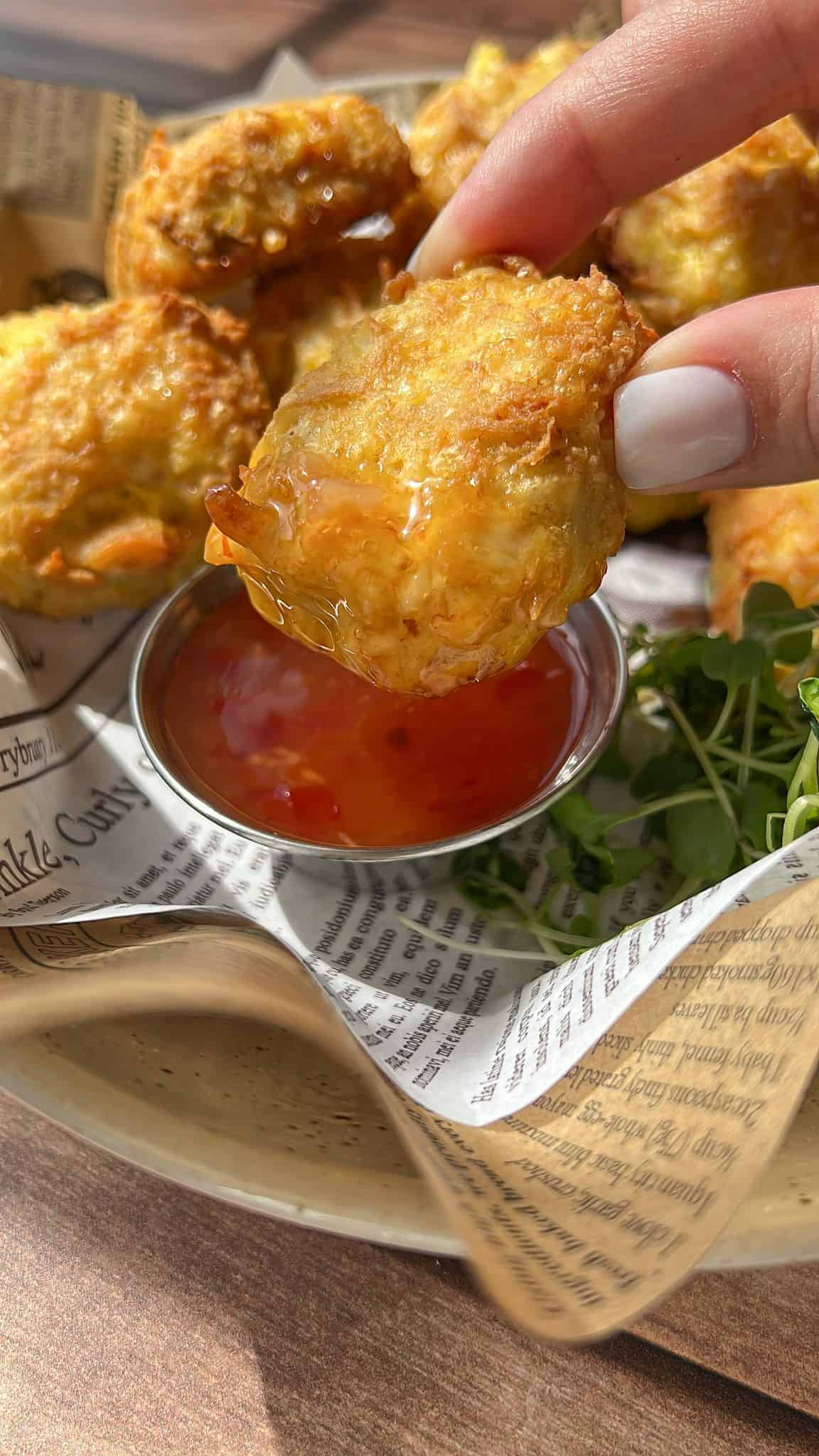 A hand holding a golden, crispy nugget dipped in orange sweet chili sauce, with more nuggets and a small dish of sauce on a newspaper-lined plate in the background.