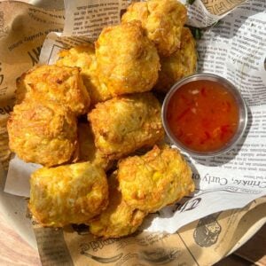 A bowl lined with newspaper-like paper holds a serving of golden, crispy fried snacks next to a small metal cup filled with orange dipping sauce. Sunlight highlights the food from above.