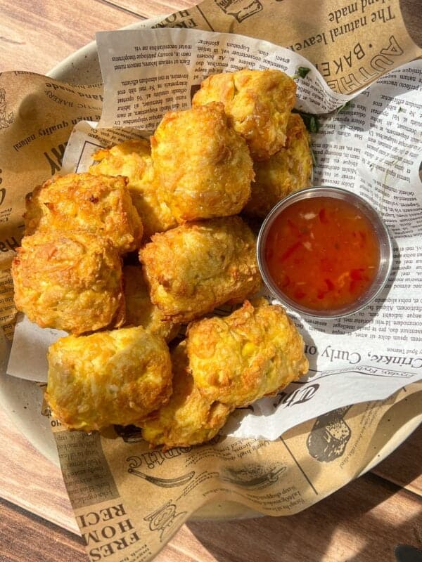 A bowl lined with newspaper-like paper holds a serving of golden, crispy fried snacks next to a small metal cup filled with orange dipping sauce. Sunlight highlights the food from above.
