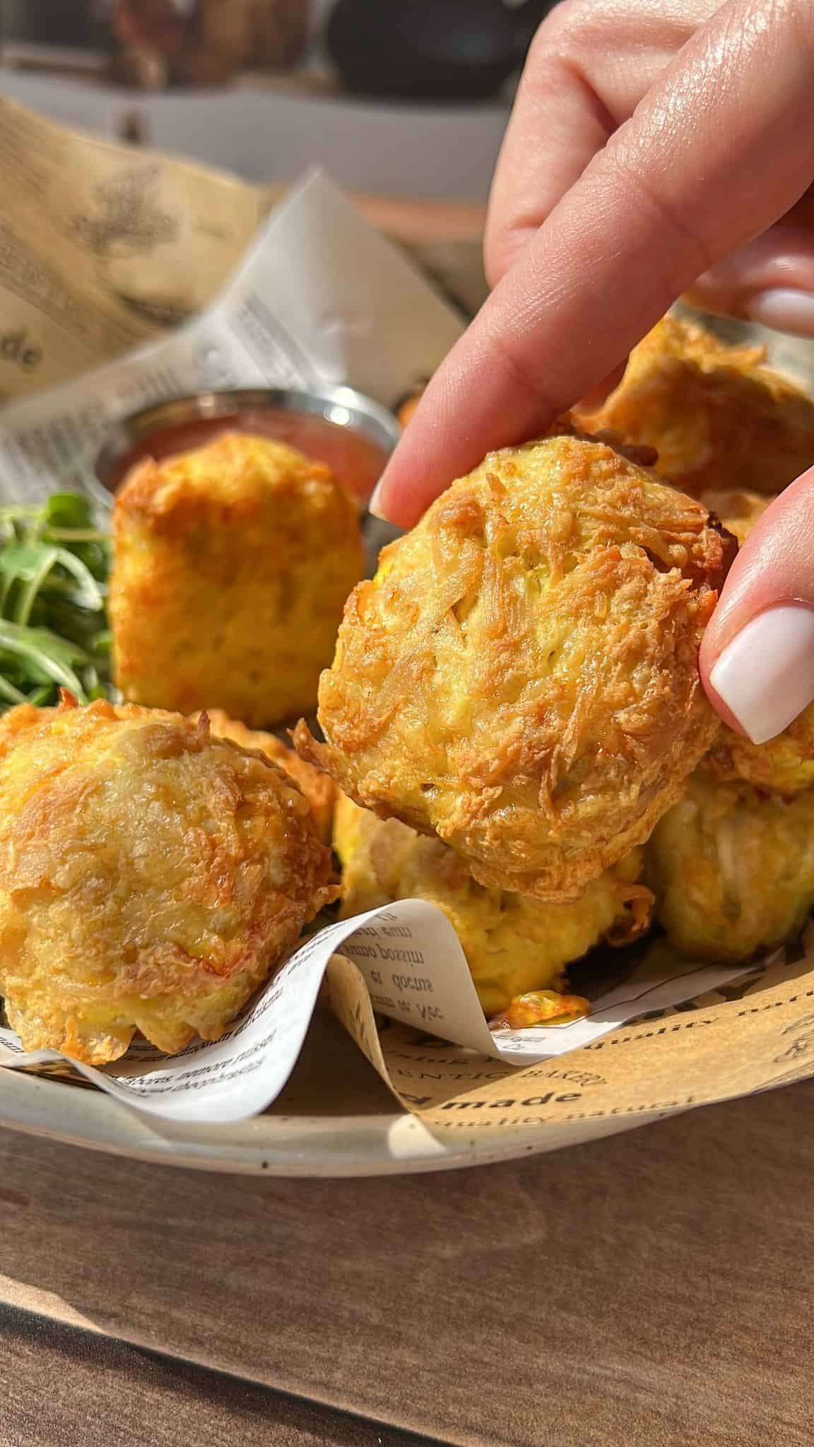 A hand with white nail polish holds a golden, crispy air fryer chicken nuggets above a bowl filled with more tater tots, a side of greens, and a cup of dipping sauce, all served on newspaper-like paper.