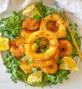 A plate of crispy, golden-brown fried onion rings rests on a bed of fresh arugula. Lemon wedges are placed around the onion rings, and a fork lies beside the plate. Bright lighting highlights the texture and colors of the dish.