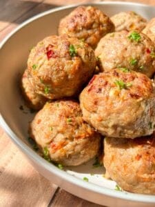 A bowl of golden-brown meatballs garnished with chopped parsley. The meatballs are visibly seasoned with herbs and spices, adding color and texture to the dish. Sunlight enhances the dish's appetizing appearance.