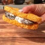 A hand holds a sandwich made with two crispy, breaded hash browns as buns, filled with cookies and cream ice cream, against a wooden table background with kitchen items blurred in the distance.