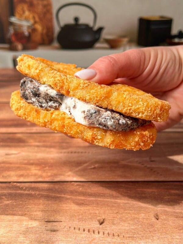 A hand holds a sandwich made with two crispy, breaded hash browns as buns, filled with cookies and cream ice cream, against a wooden table background with kitchen items blurred in the distance.