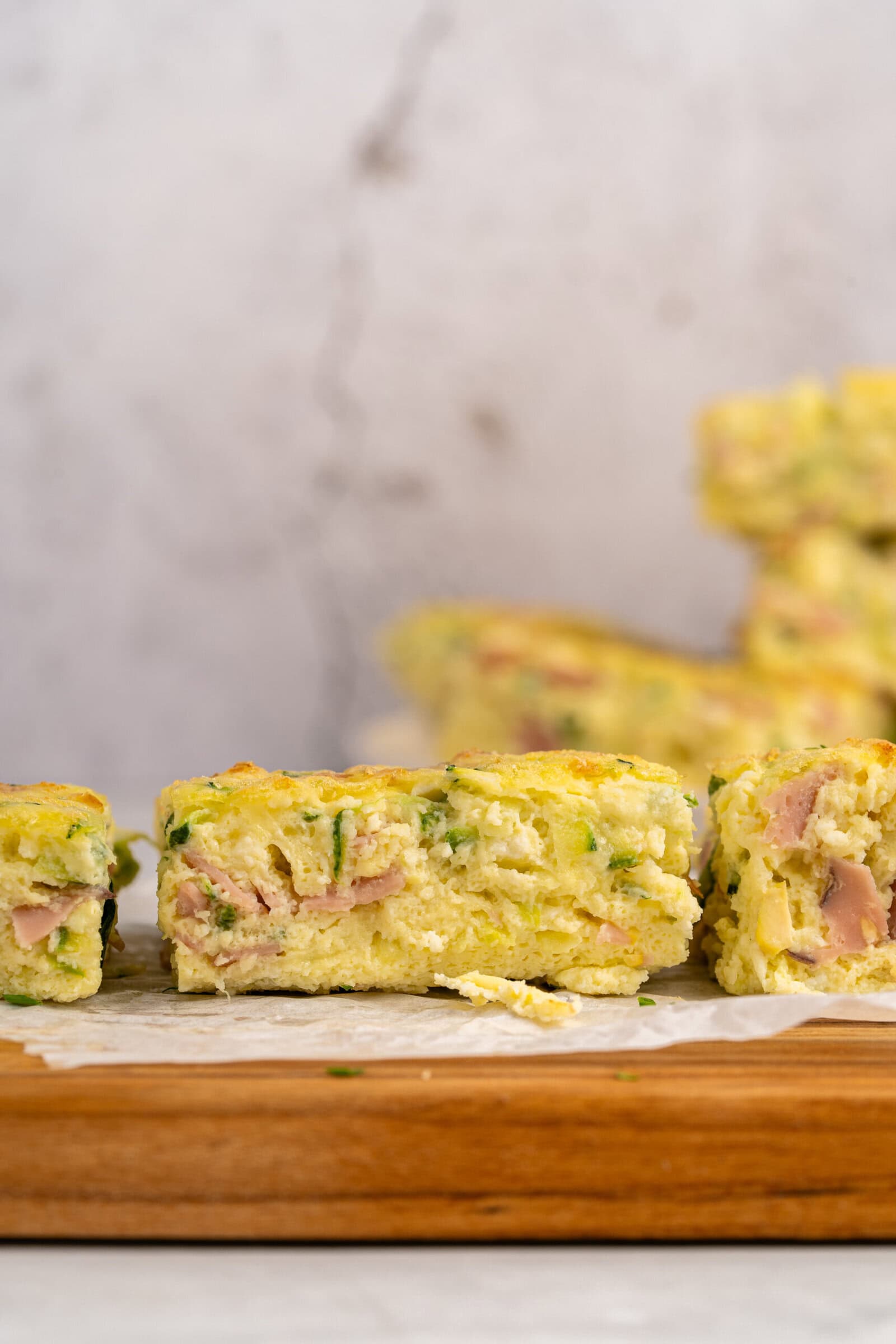 Close-up of a slice of savory egg bake with visible pieces of ham and green herbs, resting on parchment paper atop a wooden board. More pieces are blurred in the background.