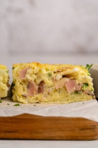 A close-up of a thick, fluffy slice of savory egg casserole with visible pieces of ham, cheese, and green herbs, resting on a wooden board lined with parchment paper.