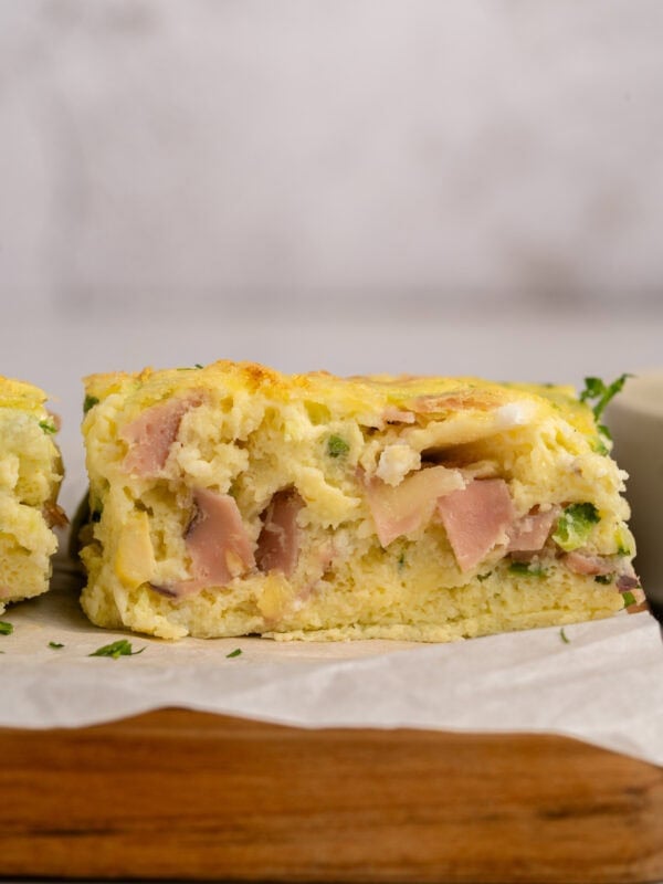 A close-up of a thick, fluffy slice of savory egg casserole with visible pieces of ham, cheese, and green herbs, resting on a wooden board lined with parchment paper.