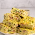 A plate stacked with thick, square slices of egg bake filled with chopped ham and green herbs, set on a light-colored surface with a blurred background.