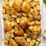 Baked Parmesan Chicken drumsticks with golden, roasted potatoes and lemon wedges, garnished with fresh herbs, arranged on parchment paper over a baking sheet. Small bowls of herbs and grated cheese are nearby.