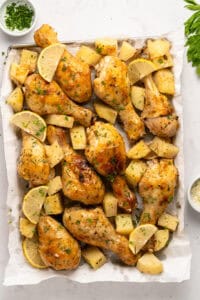 Baked Parmesan Chicken drumsticks with golden, roasted potatoes and lemon wedges, garnished with fresh herbs, arranged on parchment paper over a baking sheet. Small bowls of herbs and grated cheese are nearby.