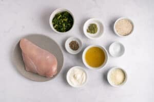 Raw chicken breast on a plate, surrounded by small bowls containing chopped spinach, pesto, rice, seasoning, broth, salt, mayonnaise, and grated cheese on a white marble surface.
