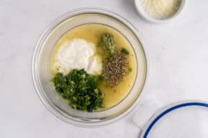 A glass bowl on a white surface containing yogurt, pesto, chopped greens, herbs, and a liquid mixture. A small bowl with grated cheese sits nearby.