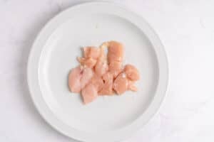 Raw, cubed pieces of chicken breast sprinkled with salt are arranged on a white plate, set against a light, marble-like background.