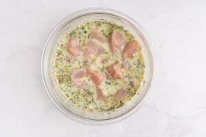 A glass bowl containing raw chicken pieces marinating in a creamy, herb-filled mixture, placed on a white marble surface.