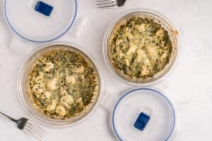 Two round glass containers filled with a cheesy, baked spinach and chicken casserole, with matching plastic lids nearby and two metal forks on a white marble surface.