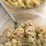 A close-up of a spoonful of rice casserole with chunks of chicken and herbs, held above a glass bowl filled with more of the same dish. Another dish of casserole is visible blurred in the background.