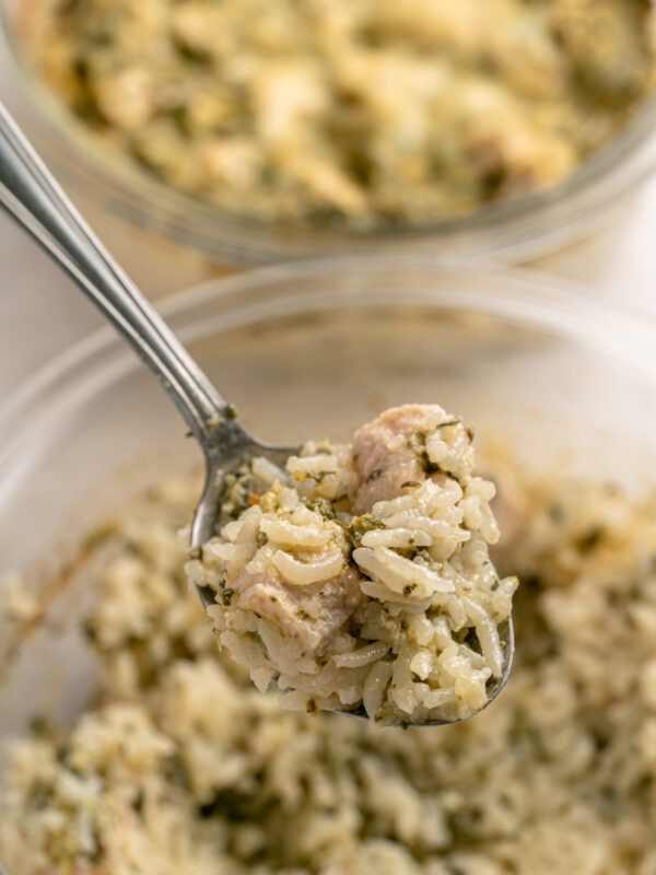 A close-up of a spoonful of rice casserole with chunks of chicken and herbs, held above a glass bowl filled with more of the same dish. Another dish of casserole is visible blurred in the background.