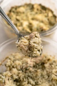 A close-up of a spoonful of rice with herbs and pieces of chicken, held above a bowl filled with the same mixture. Another similar bowl is visible in the background.