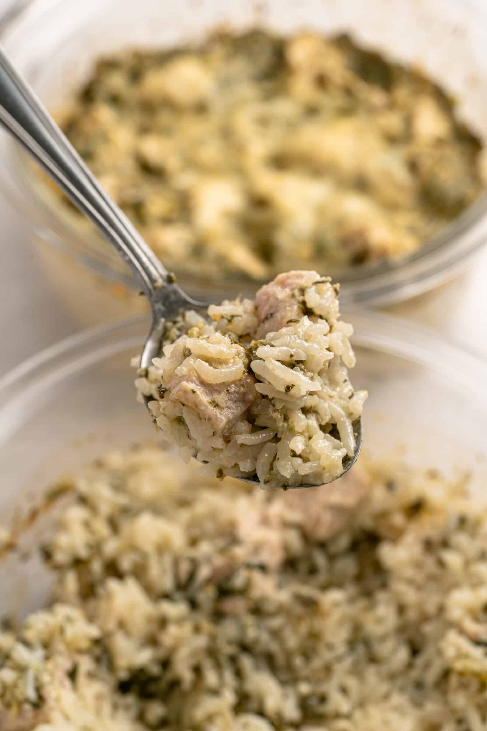 A close-up of a spoonful of rice with herbs and pieces of chicken, held above a bowl filled with the same mixture. Another similar bowl is visible in the background. - 3