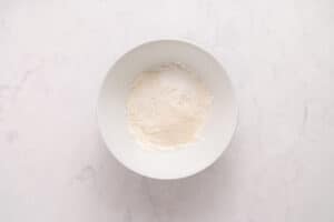 A white bowl containing a small mound of flour sits on a light marble surface.