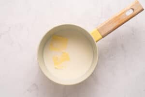 A saucepan with a wooden handle contains a creamy liquid and two partially melted pats of butter on a light marble surface.