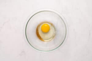 A clear glass bowl on a white surface contains a raw egg with an intact yolk and some brown seasoning or liquid around the egg white.