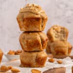 Three banana muffins are stacked on a wooden board, with the top muffin frosted and sprinkled with chopped almonds. Additional muffins and whole almonds are in the background on a light, textured surface.