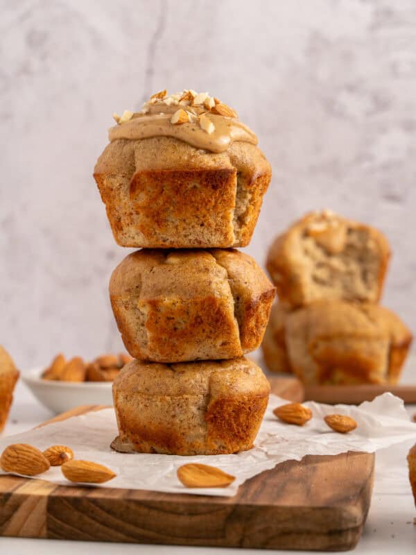 Three banana muffins are stacked on a wooden board, with the top muffin frosted and sprinkled with chopped almonds. Additional muffins and whole almonds are in the background on a light, textured surface.