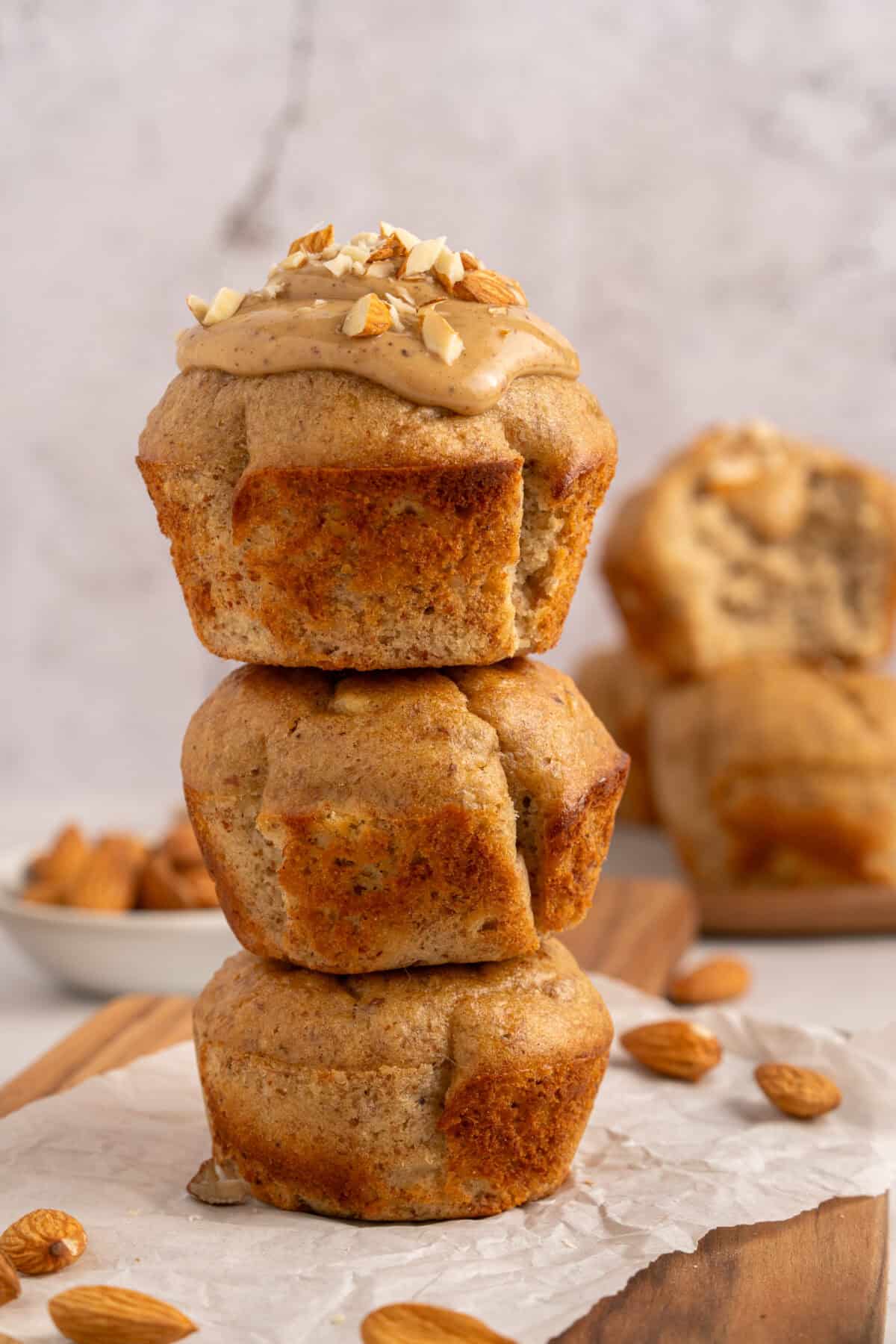 Three almond muffins are stacked on top of each other. The top muffin is topped with almond butter and chopped almonds. More muffins and a small bowl of almonds are blurred in the background.