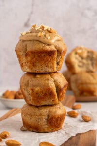 Three almond muffins are stacked on top of each other. The top muffin is topped with almond butter and chopped almonds. More muffins and a small bowl of almonds are blurred in the background.