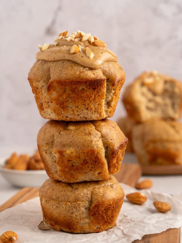 Three almond muffins are stacked on top of each other. The top muffin is topped with almond butter and chopped almonds. More muffins and a small bowl of almonds are blurred in the background.