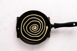A nonstick frying pan on a white surface contains pancake batter piped in a spiral pattern, ready to be cooked.