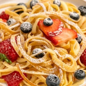 A plate of fettuccine pasta topped with fresh strawberries, blueberries, and a dusting of powdered sugar.