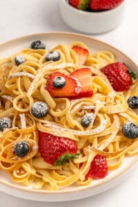 A plate of fettuccine pasta topped with fresh strawberries, blueberries, and a dusting of powdered sugar, with a bowl of strawberries in the background.
