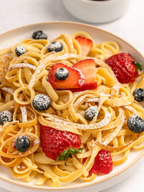 A plate of fettuccine pasta topped with fresh strawberries, blueberries, and a dusting of powdered sugar, with a bowl of strawberries in the background.