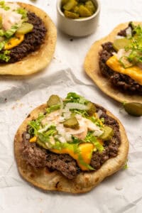 Three open-faced cheeseburger flatbreads topped with melted cheese, lettuce, chopped onions, sliced pickles, and sauce, on parchment paper. A small bowl of extra pickles is in the background.