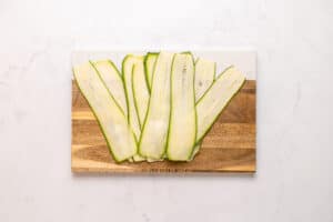 Thin slices of zucchini neatly arranged on a wooden cutting board, placed on a white marble surface.