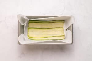 A loaf pan lined with parchment paper and layered with thin, overlapping slices of zucchini on the bottom.