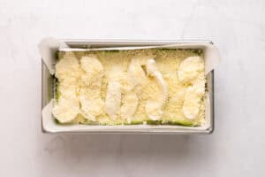 A loaf pan lined with parchment paper contains an unbaked casserole, topped with shredded cheese and dollops of a creamy white mixture on a light marble surface.