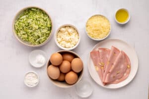 Bowls and plates with ingredients on a white surface: grated zucchini, ricotta cheese, shredded cheese, four eggs, slices of mortadella, flour, baking powder, salt, and a small bowl of olive oil.