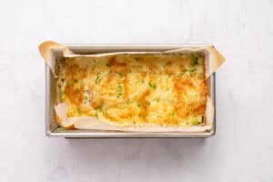 A golden-brown cheesy casserole topped with herbs sits in a parchment-lined loaf pan on a white marble surface.