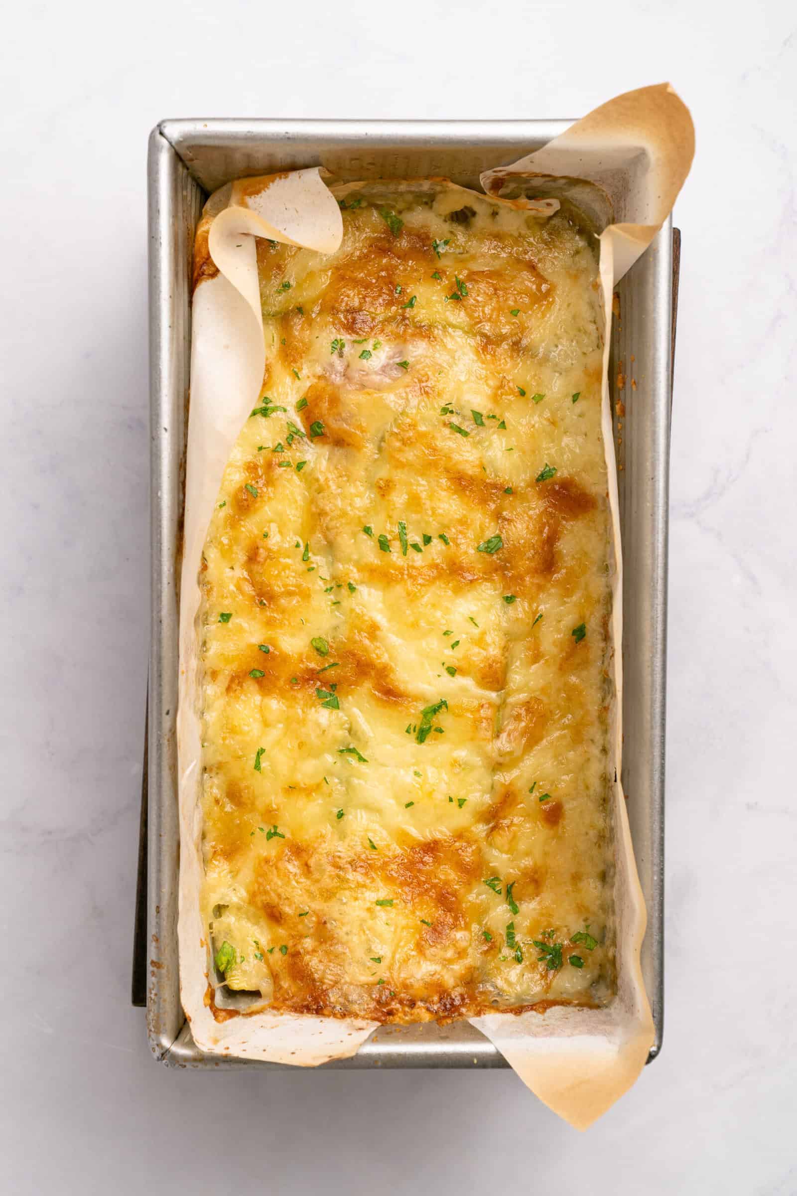 A loaf pan lined with parchment paper holds a golden-brown, cheesy baked casserole topped with chopped herbs, resting on a light marble surface.