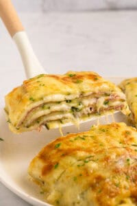 A close-up of a cheesy, layered baked dish being lifted from a plate, revealing layers of melted cheese, ham, and sauce, topped with golden, bubbly cheese and sprinkled with herbs.