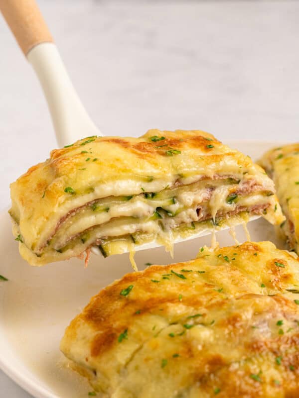 A close-up of a cheesy, layered baked dish being lifted from a plate, revealing layers of melted cheese, ham, and sauce, topped with golden, bubbly cheese and sprinkled with herbs.