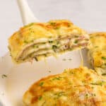 A cheesy slice of baked lasagna with herbs is lifted from a white plate, showing layers of pasta, melted cheese, and filling. Another portion remains on the plate in the background.