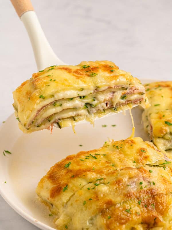 A cheesy slice of baked lasagna with herbs is lifted from a white plate, showing layers of pasta, melted cheese, and filling. Another portion remains on the plate in the background.