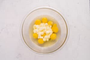A glass bowl containing several raw eggs and a white, crumbly ingredient, possibly ricotta cheese, sits on a light marble surface.